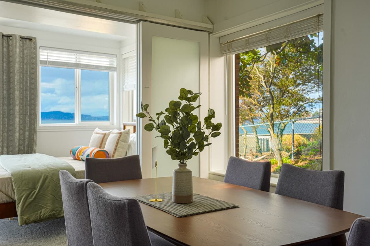 Airy dining area features a sleek wooden table and plush seating, surrounded by large windows that fill the space with natural light and coastal views. A sliding door opens to a cozy bedroom, blending comfort with scenic tranquility.