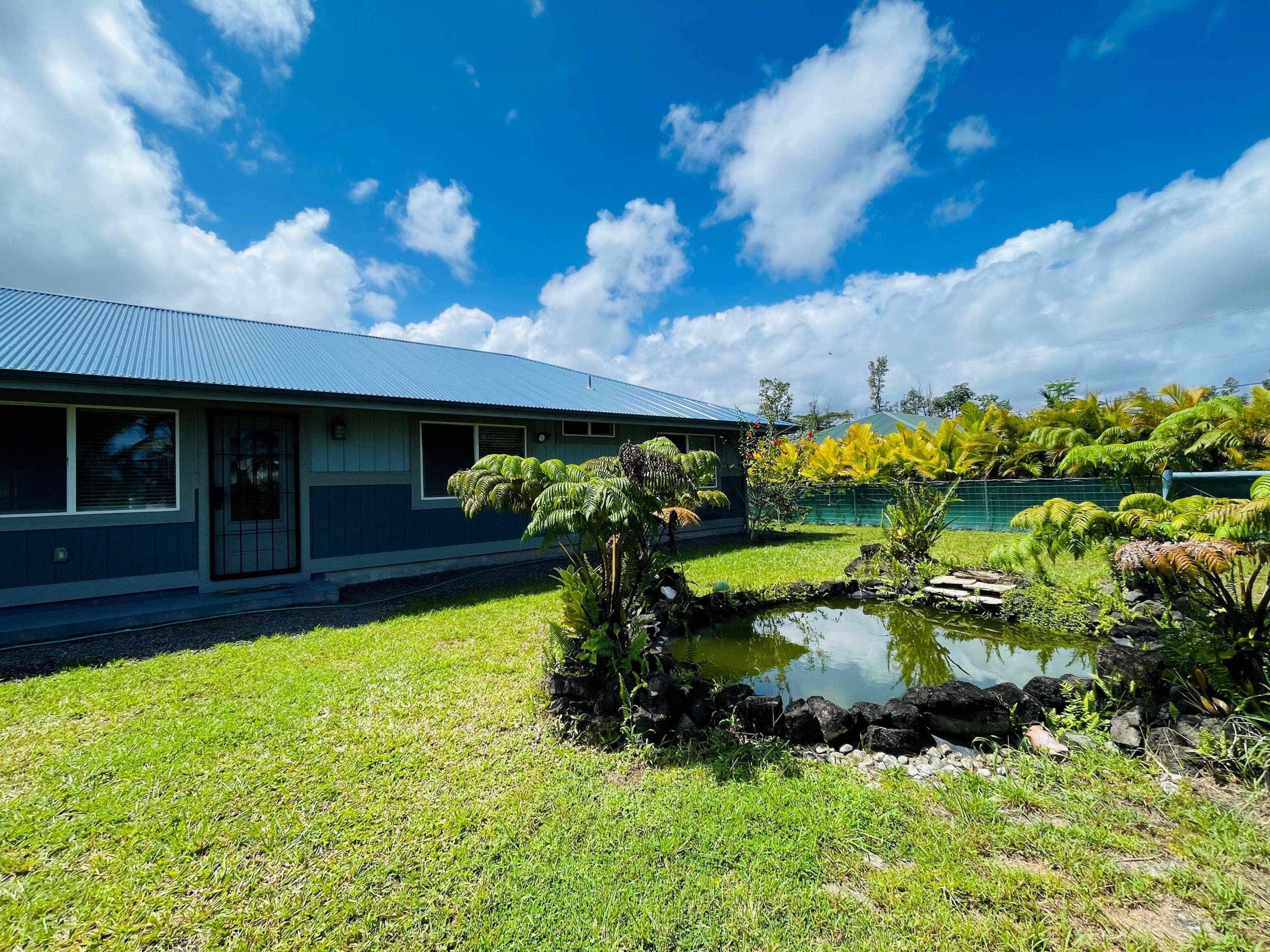 Charming single-story home with a metal roof, surrounded by lush tropical landscaping and featuring a serene private pond in the front yard.