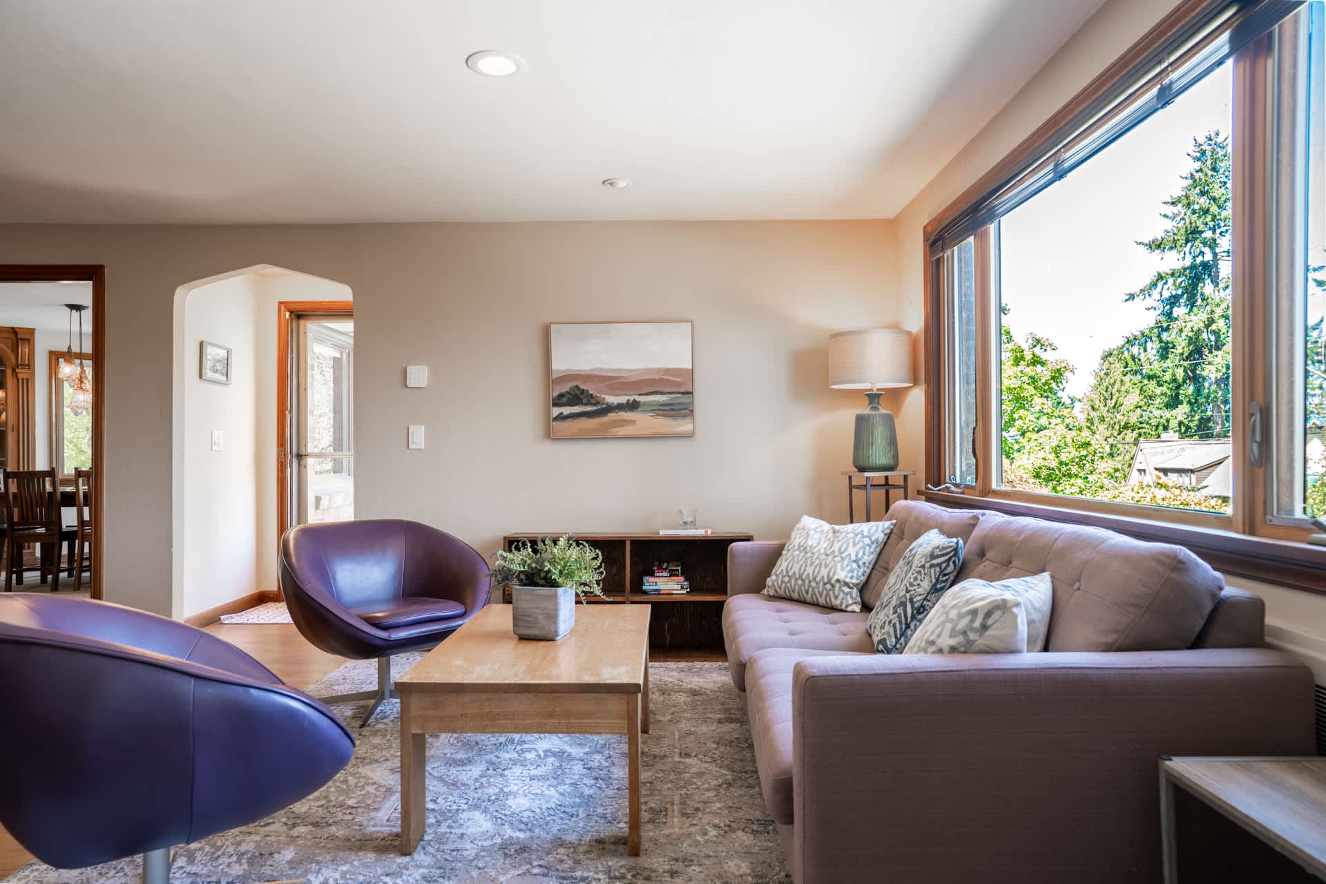 This bright, inviting living room offers a warm, relaxing experience with its large sunlit windows, cozy seating, and serene view of greenery outside.